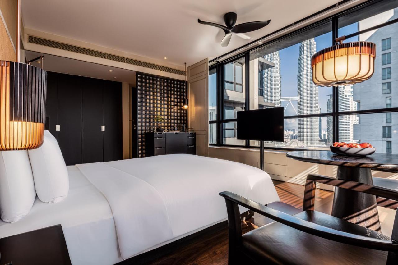 The Ruma Hotel And Residences Куала Лумпур Екстериор снимка The photo shows a modern hotel room featuring a large bed with white linens. There's a ceiling fan above and large windows letting in natural light, providing a view of a cityscape outside. To one side, there's a small seating area with a round table